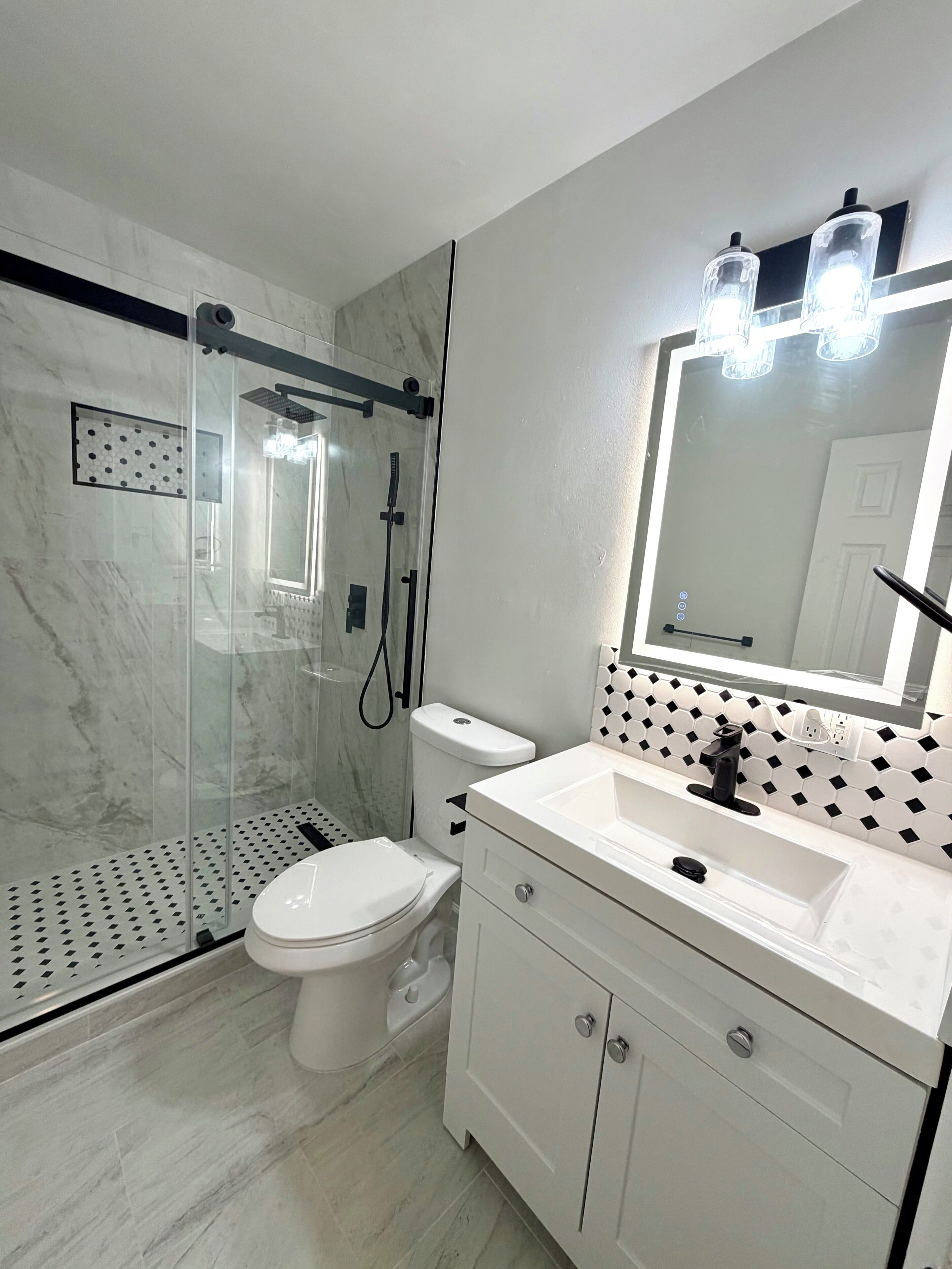 Une salle de bain moderne avec une douche à parois de verre, des toilettes, un lavabo avec un miroir et un mur recouvert de carreaux.