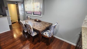 A dining area with a wooden table and floral-patterned chairs, a window with floral curtains, and a kitchen in the background.