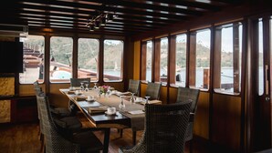 Un coin repas avec une longue table dressée pour quatre personnes, entourée de chaises en osier, de grandes fenêtres offrant une vue sur les collines et un plafond en bois.