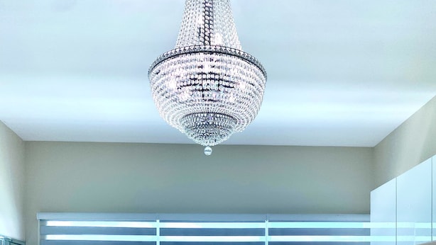 Un salon moderne avec un canapé blanc, deux coussins bleus, une table basse en verre et un lustre. De grandes fenêtres offrent une vue sur l’océan.