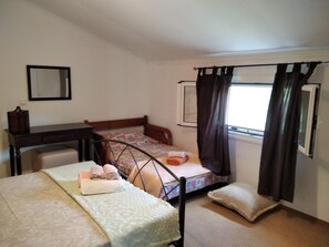 Een kleine kamer met een enkel bed, een houten hoofdbord, nachtkastje, raam met gordijnen en een spiegel aan de muur.