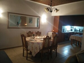 Een eetruimte met een ronde tafel voor zes personen, omringd door houten stoelen. Een kroonluchter hangt aan het plafond en een grote spiegel is aan de muur boven de tafel bevestigd.