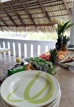 Un espacio al aire libre cubierto con techo de paja, una mesa con frutas y un dispensador de bebida verde, y platos blancos apilados con diseños verdes.
