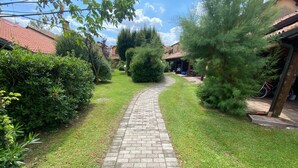 A paved pathway leading through a well-maintained garden with various shrubs and trees, surrounded by residential buildings. - Beautiful and Quiet Villa In Residence In The Po Delta Oasis (Porto Viro)
