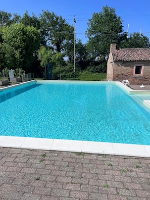 Una piscina rettangolare con acqua cristallina, circondata da una zona pavimentata e un edificio in mattoni con tetto in tegole.