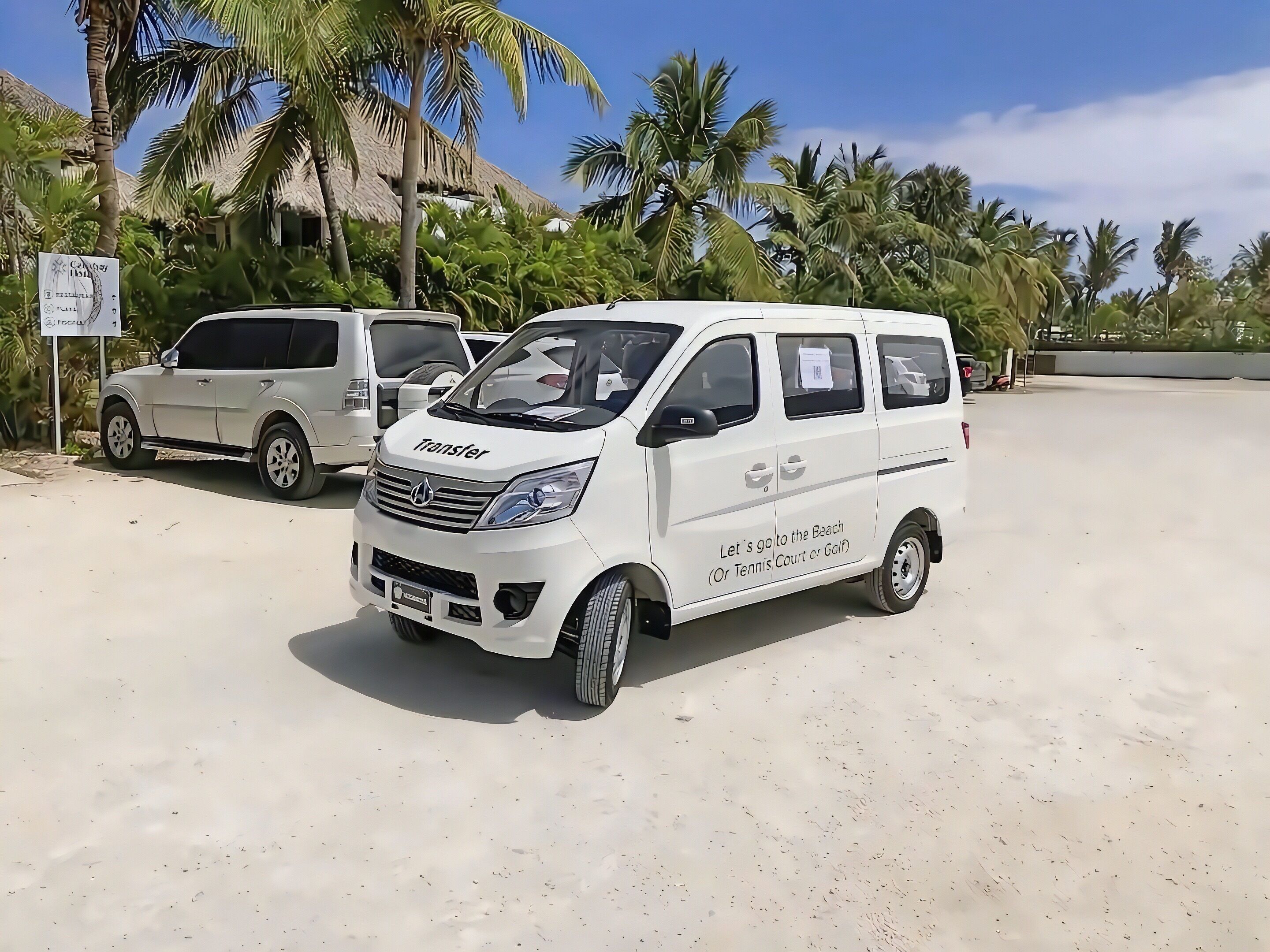 A white van with 'Transfer' and 'Let's go to the Beach (Or Tennis Court or Golf)' text, parked on a sandy surface with palm trees and a thatched-roof building in the background.