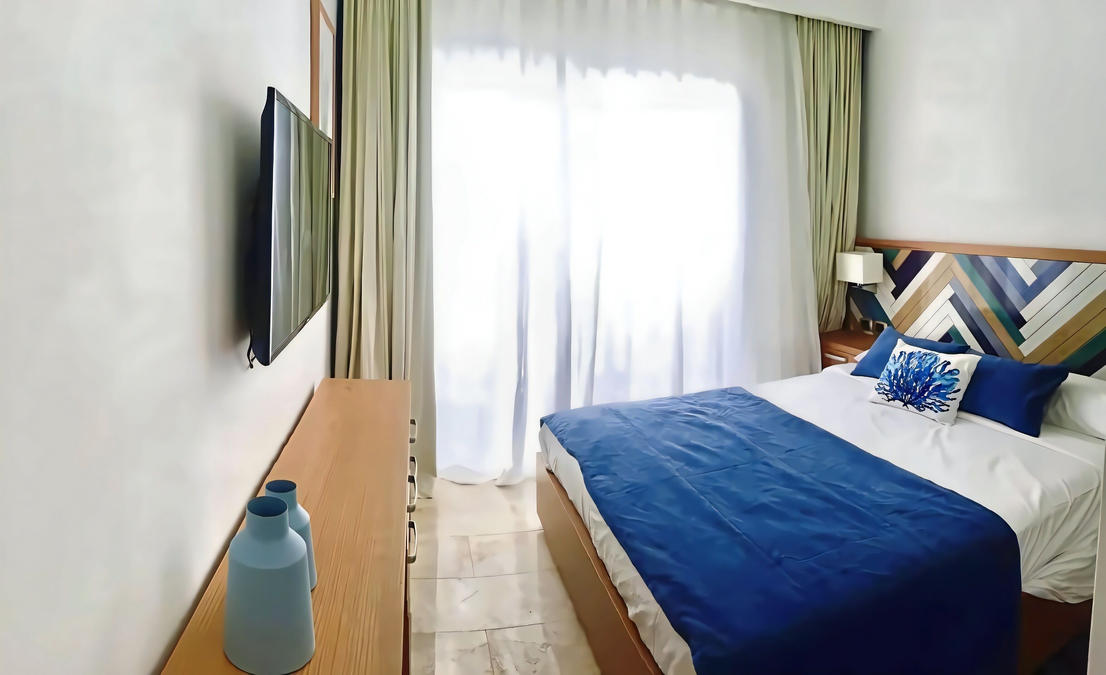 Ein Hotelzimmer mit Bett, Fernseher, Nachttisch, Lampe und Blick ins Freie.