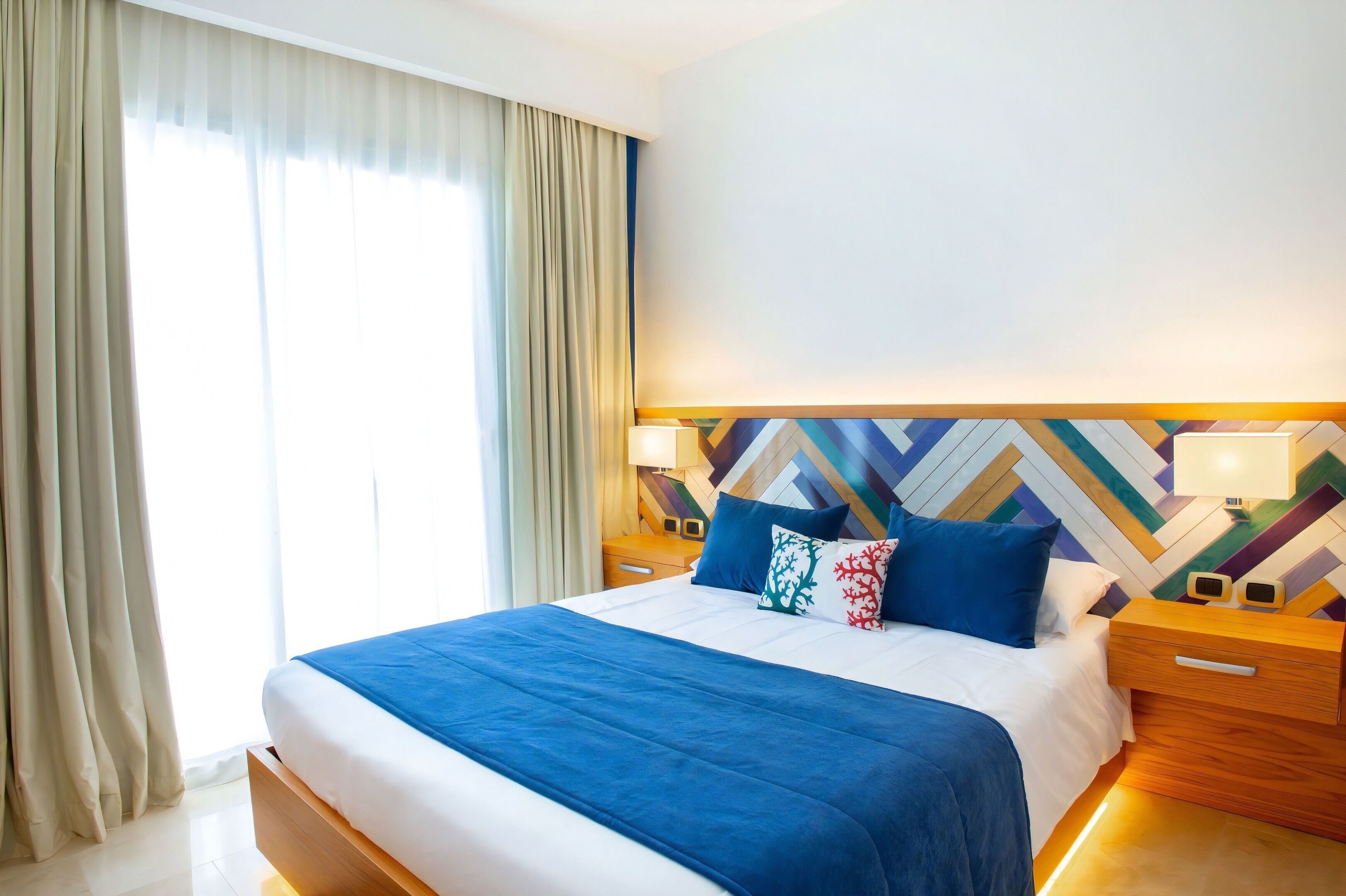 Habitación de hotel con una cama grande, dos mesitas de noche, un diseño de pared moderno y un ventanal con cortinas.