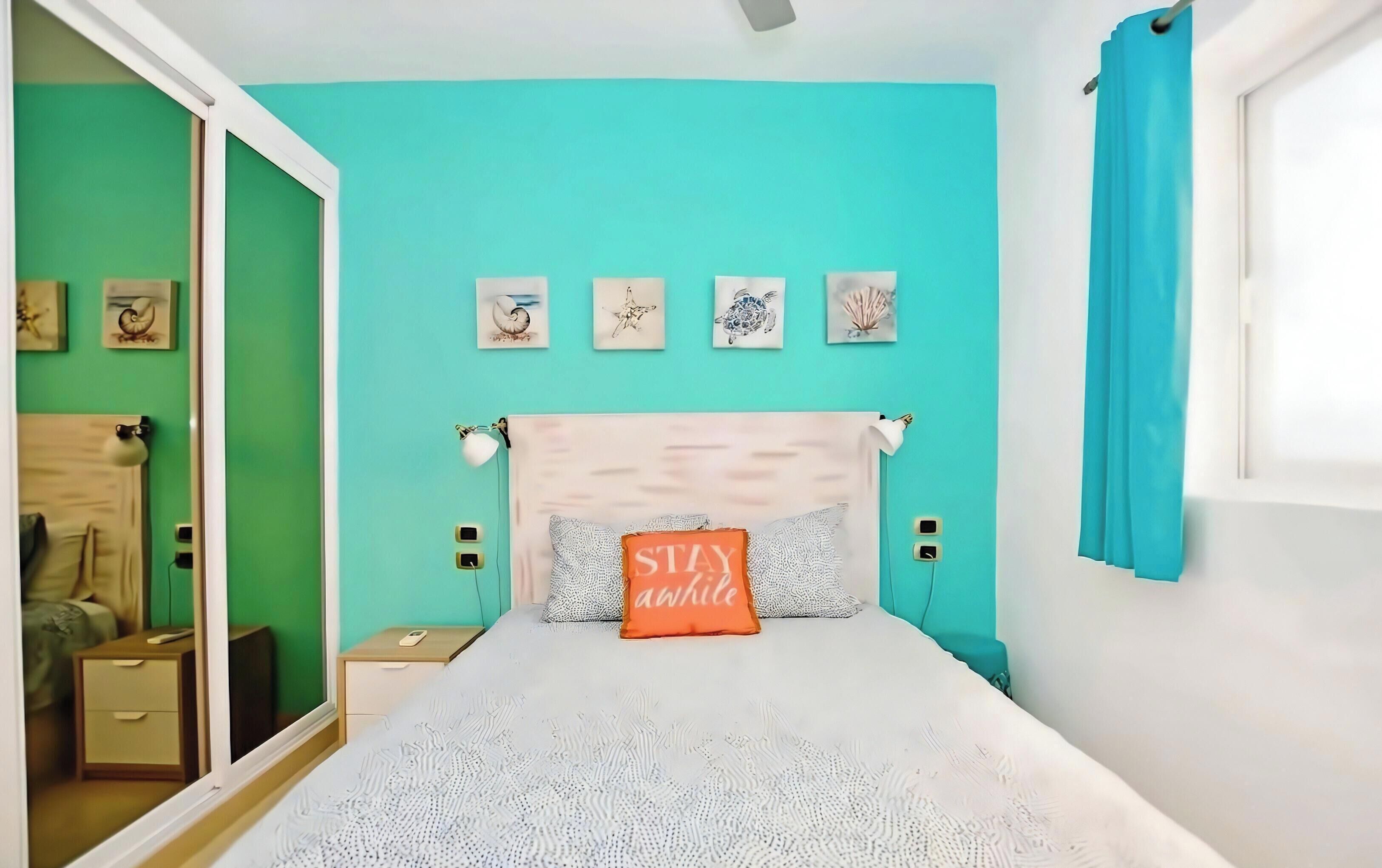Une chambre avec un mur de couleur turquoise, un lit avec une tête de lit blanche et un coussin orné de la mention « Restez un peu ».