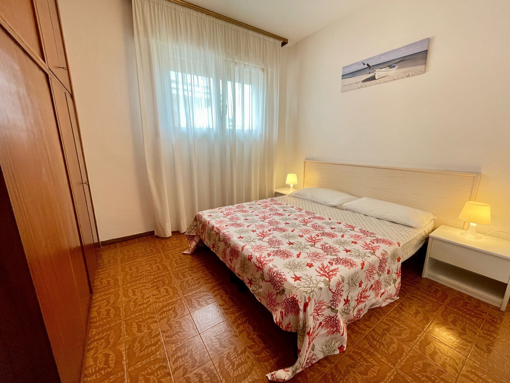 A bedroom with a bed, two bedside tables, a nightstand, a window with curtains, and a painting on the wall.