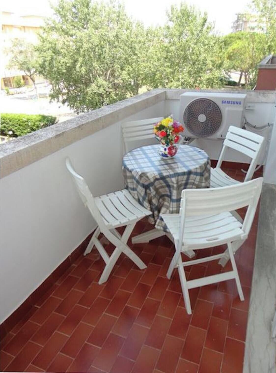A small outdoor balcony with a round table and two white chairs, a flower vase, and a wall-mounted air conditioning unit.