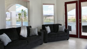 Una sala de estar con dos sofás oscuros, una manta blanca y almohadones estampados. Amplias ventanas con vistas a palmeras y montañas.