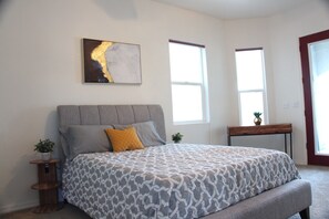A bedroom with a large bed, a headboard, two windows, a bedside table with a plant, and a painting on the wall. - On River Time!  (Bullhead City)