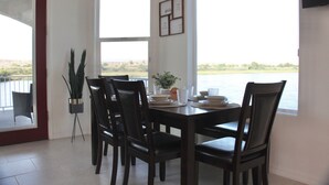 Un comedor con una mesa dispuesta para cuatro personas, rodeada de sillas, y vistas a un cuerpo de agua y al paisaje a través de amplios ventanales.