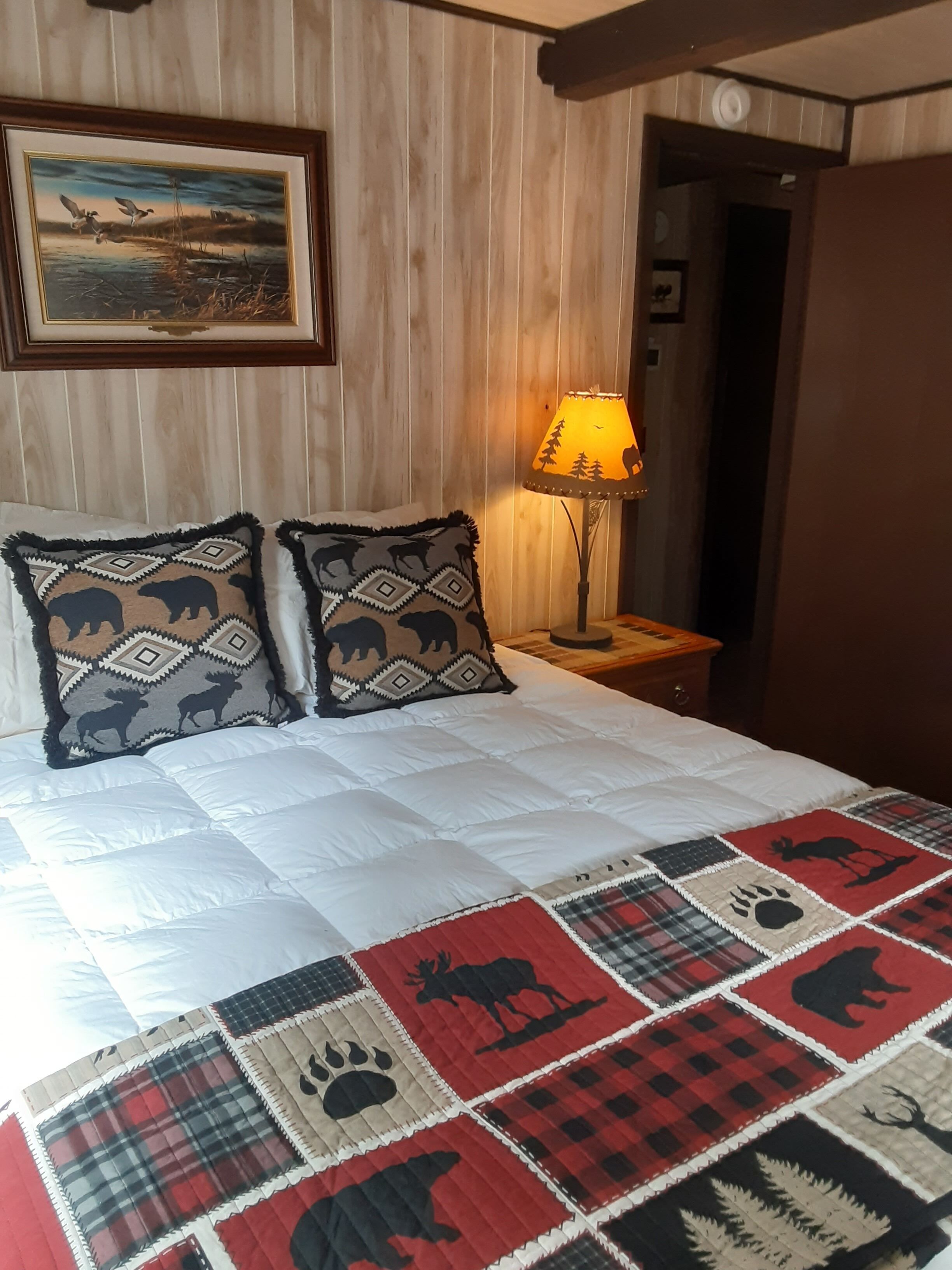 Un dormitorio con colcha acolchada estampada con motivos de animales, cabecero de madera, un cuadro enmarcado en la pared y una lámpara de noche.