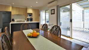 A dining area with a wooden table, chairs, and a fruit bowl. There is a kitchen in the background with wooden cabinets and appliances. - Fermoy Cottage (Yarra Ranges)