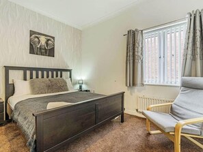 Un dormitorio con cama, una silla, una mesita de noche, una lámpara y una ventana con cortinas.