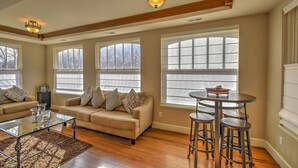 A spacious living room with a beige sofa, patterned cushions, a glass coffee table, and a round bar stool set. Large windows with blinds allow natural light.