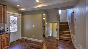 A spacious kitchen with wooden cabinets and a large window, a hallway with a wooden staircase, and a door leading to another room.