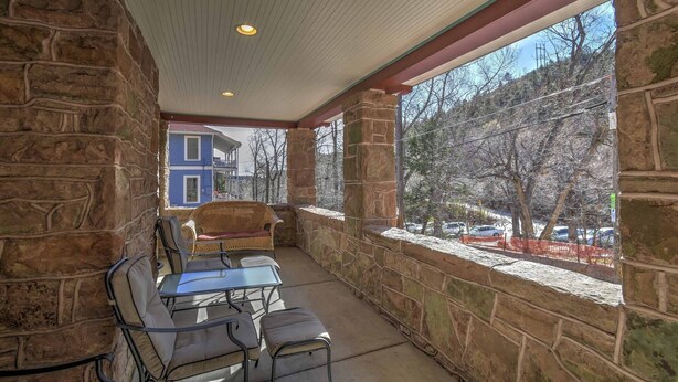A covered patio with stone walls, a wicker chair, a glass table, and a view of a blue house and trees.