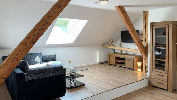 Un dormitorio tipo loft moderno con techo inclinado, techo de cristal, un sofá oscuro, un mueble de madera y una pequeña mesa con una vela.