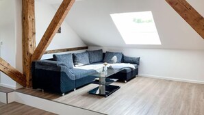 Un dormitorio tipo loft moderno con techo inclinado, vigas de madera a la vista, un ventanal grande, un sofá modular y una mesa de centro de vidrio.