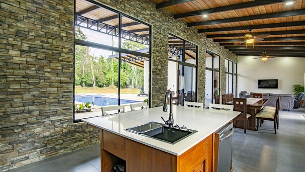Une cuisine moderne dotée d’un grand îlot central, de murs en pierre et de grandes fenêtres donnant sur un jardin avec piscine.