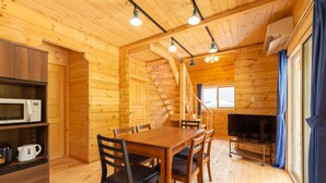 A wooden cabin interior with a dining table, chairs, a kitchen area with a microwave, and a fireplace.