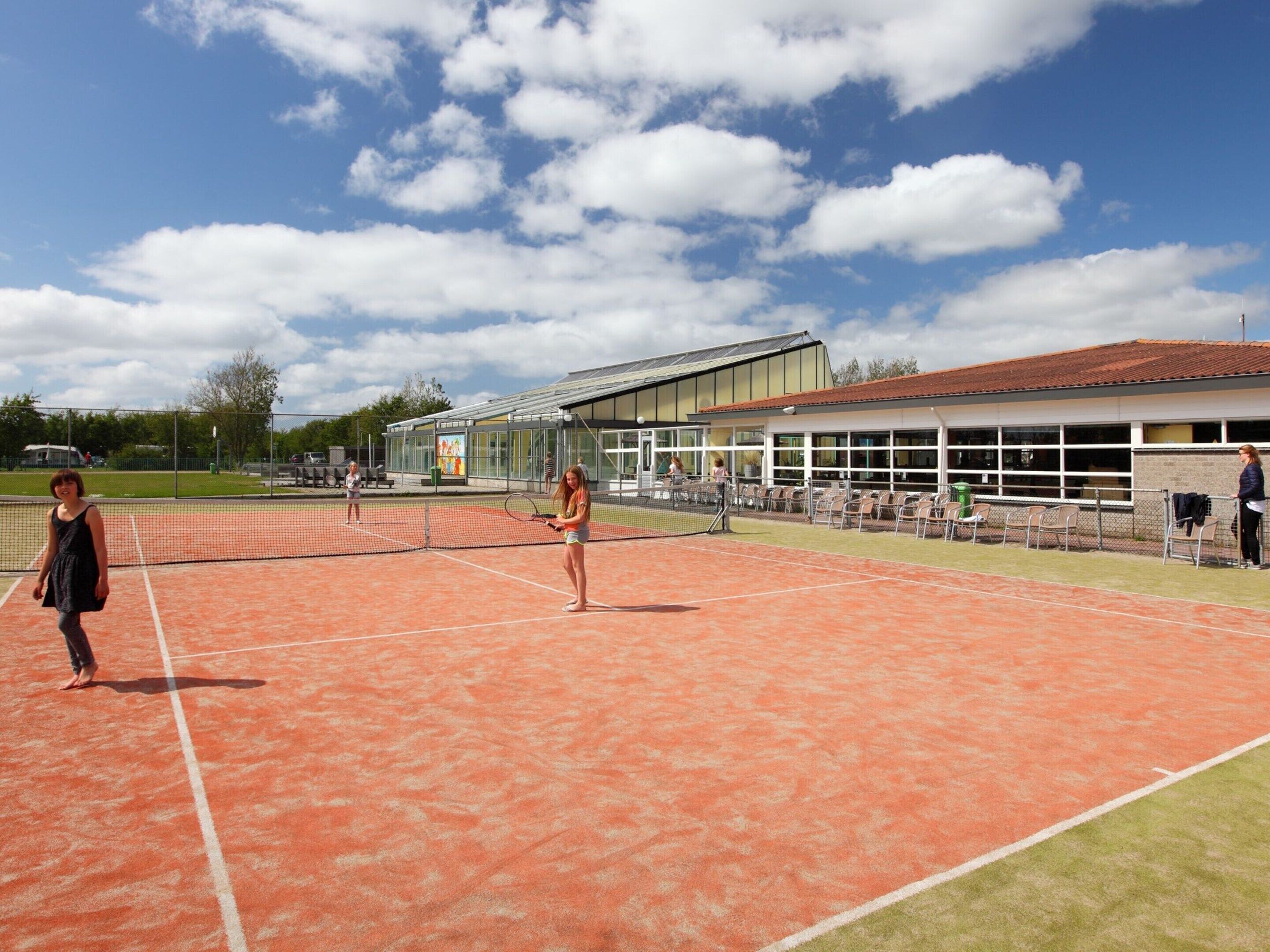 An outdoor tennis court with a net, a building with large windows, and a covered seating area.