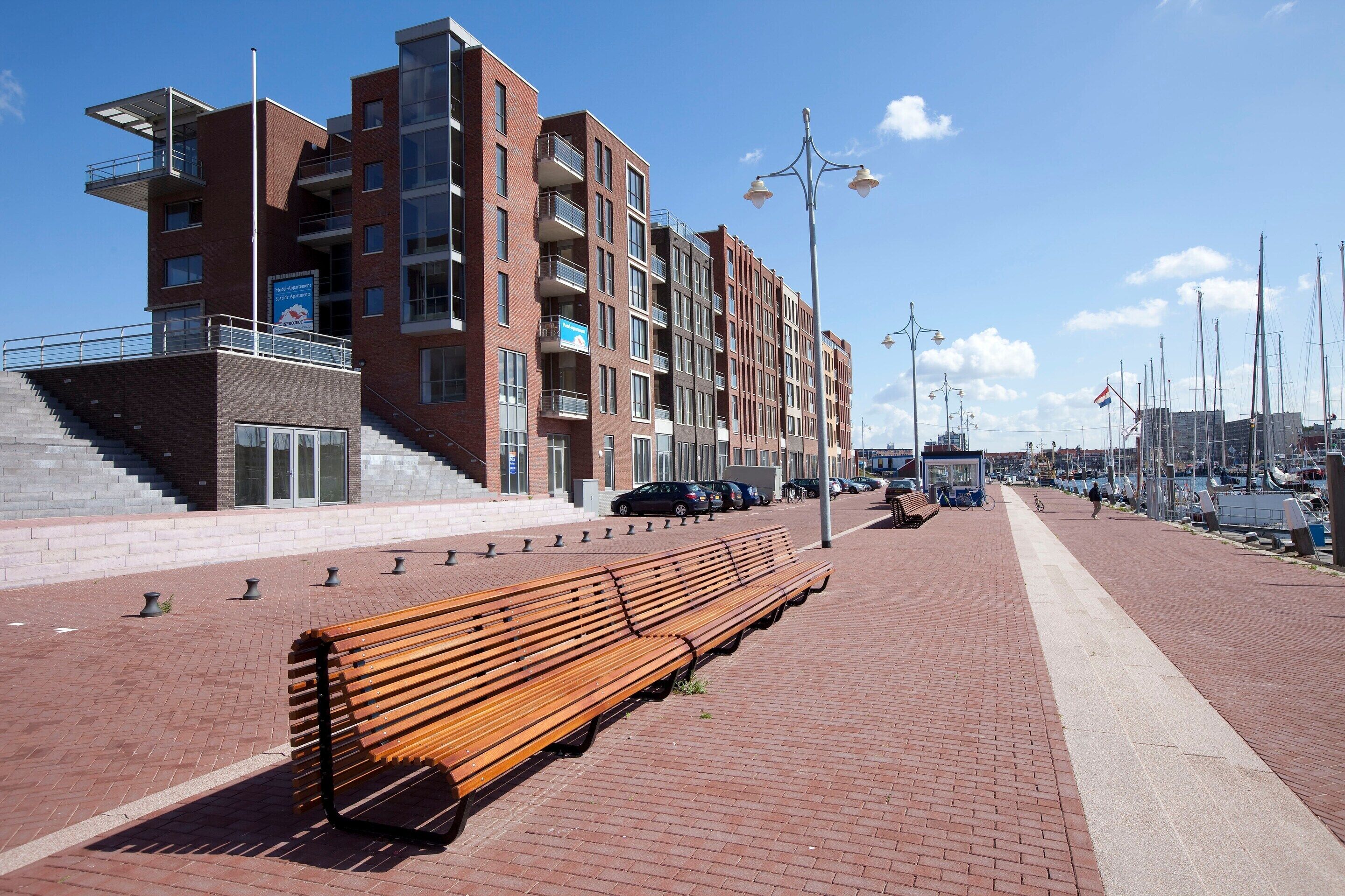 Une promenade en bord de mer avec un banc en bois, un bâtiment moderne en briques doté de balcons, et une rangée de voitures garées.