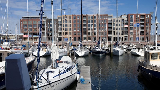 Marina com vários barcos atracados, um cais de madeira e um edifício com vários andares ao fundo.