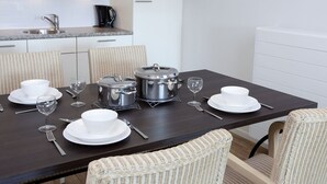 Un comedor con una mesa de madera oscura dispuesta para cuatro personas, platos, tazones y copas de vino blancos, y sillas de mimbre.