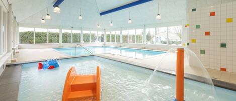 En indendørs swimmingpool med rutsjebane, et vandlegeområde og udsigt til grøn vegetation.