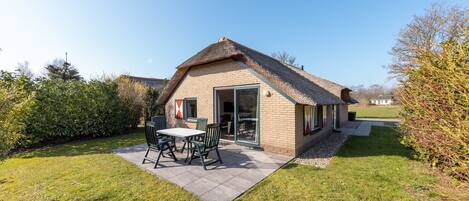 Een huis met rieten dak, een terras met tafel en stoelen, en een tuin.