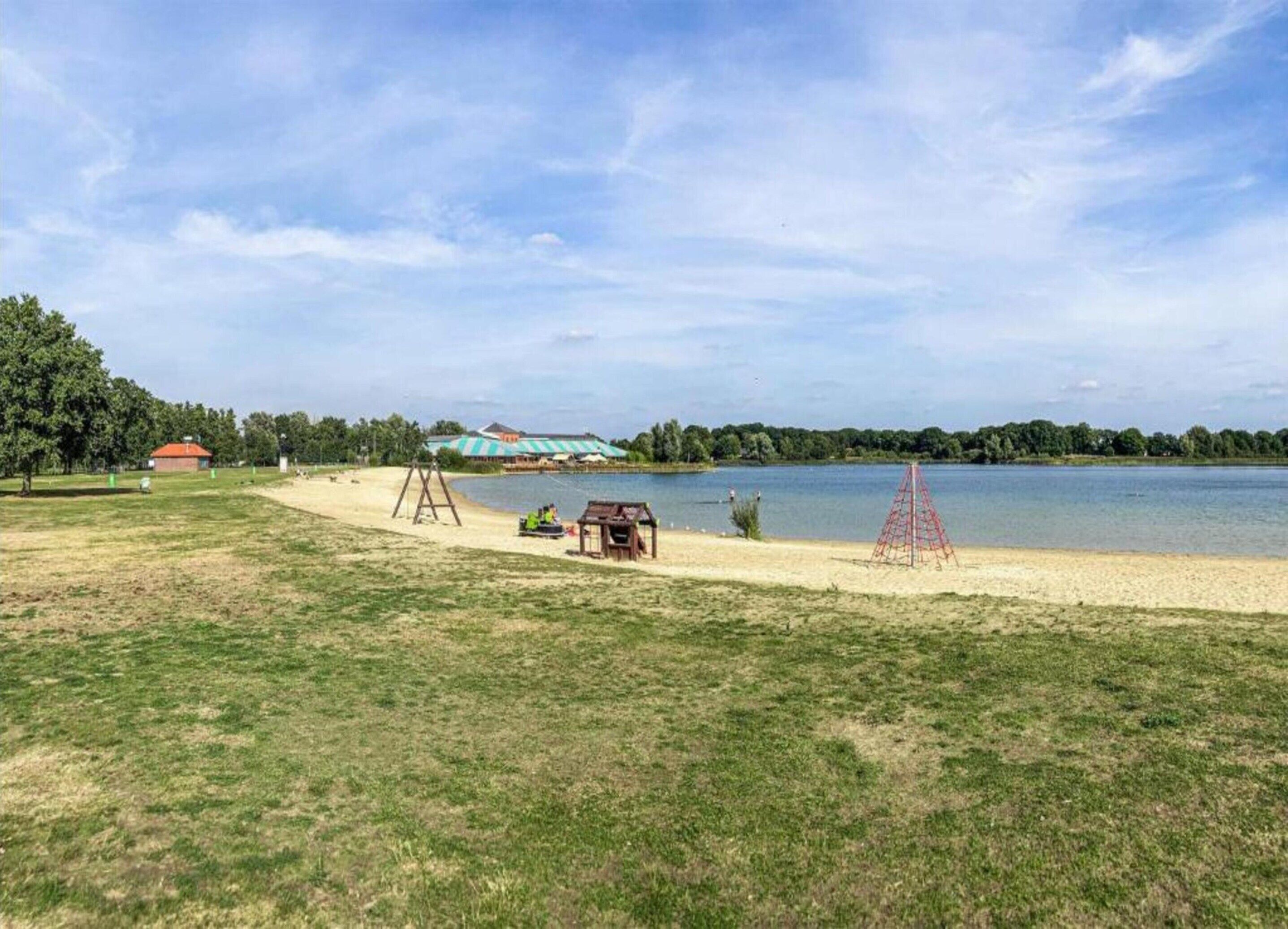 A lakeside area with a grassy field, a swing set, a small wooden structure, and a red climbing frame.
