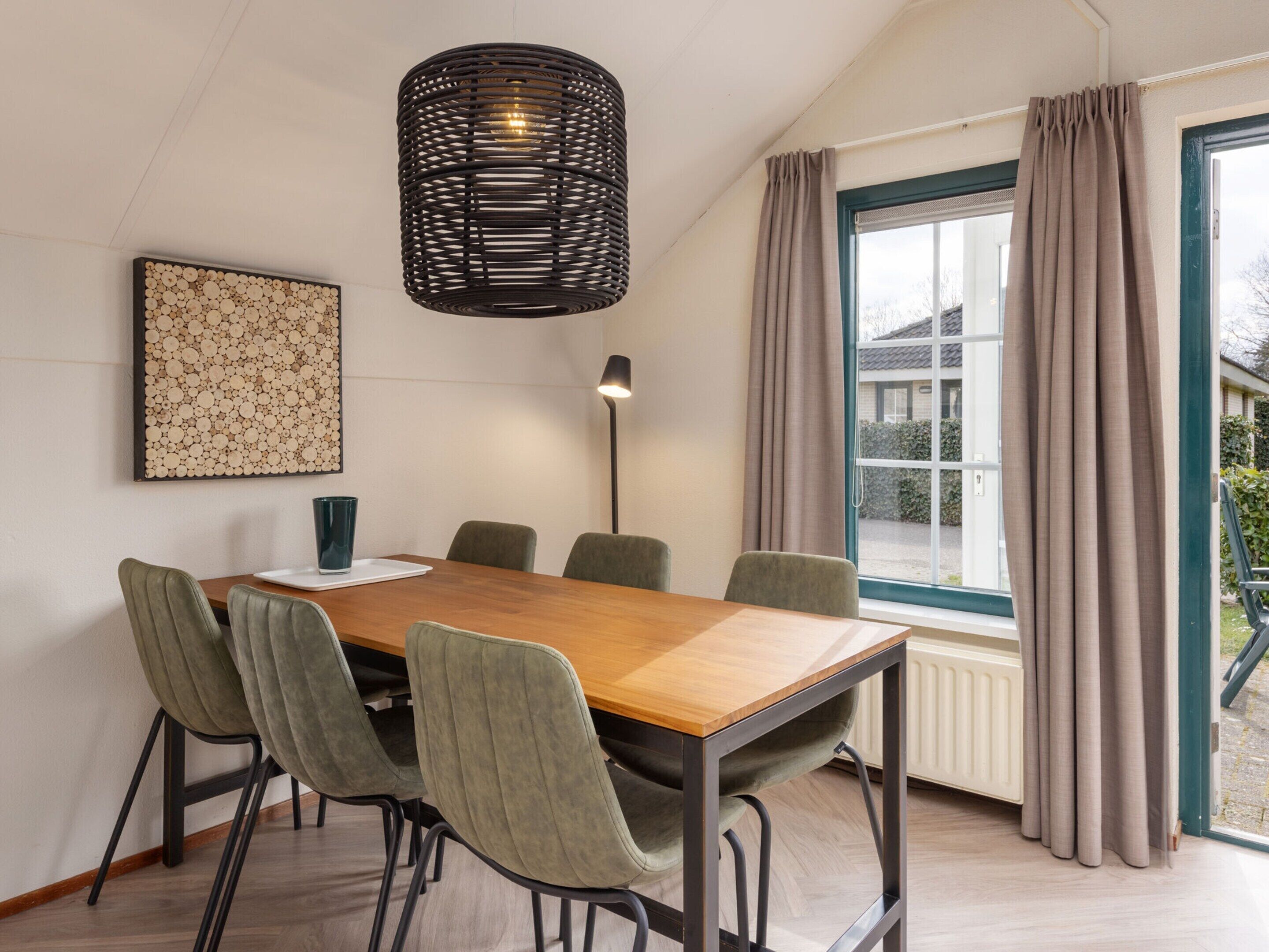 A conference room with a wooden table, six chairs, a black woven pendant light, a floor lamp, a window with curtains, and a framed abstract art piece on the wall.