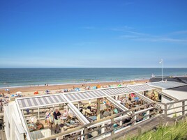 A beachside restaurant with outdoor seating overlooking the ocean, a clear sky, and a sandy beach in the background.