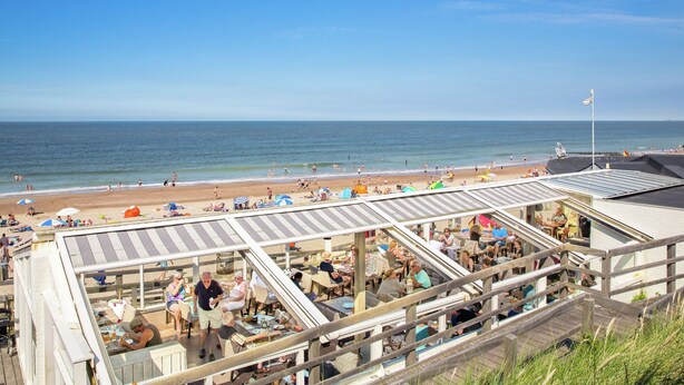 Ein Restaurant direkt am Strand mit Außenbereich und Meerblick, einem klaren Himmel und einem Sandstrand im Hintergrund.