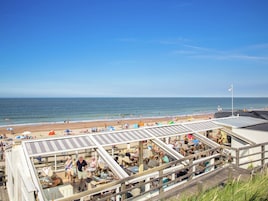 A beachside restaurant with outdoor seating overlooking the ocean, a clear sky, and a sandy beach in the background.