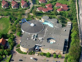 An aerial view of a building with multiple cylindrical structures, a large circular dome, and a parking area.