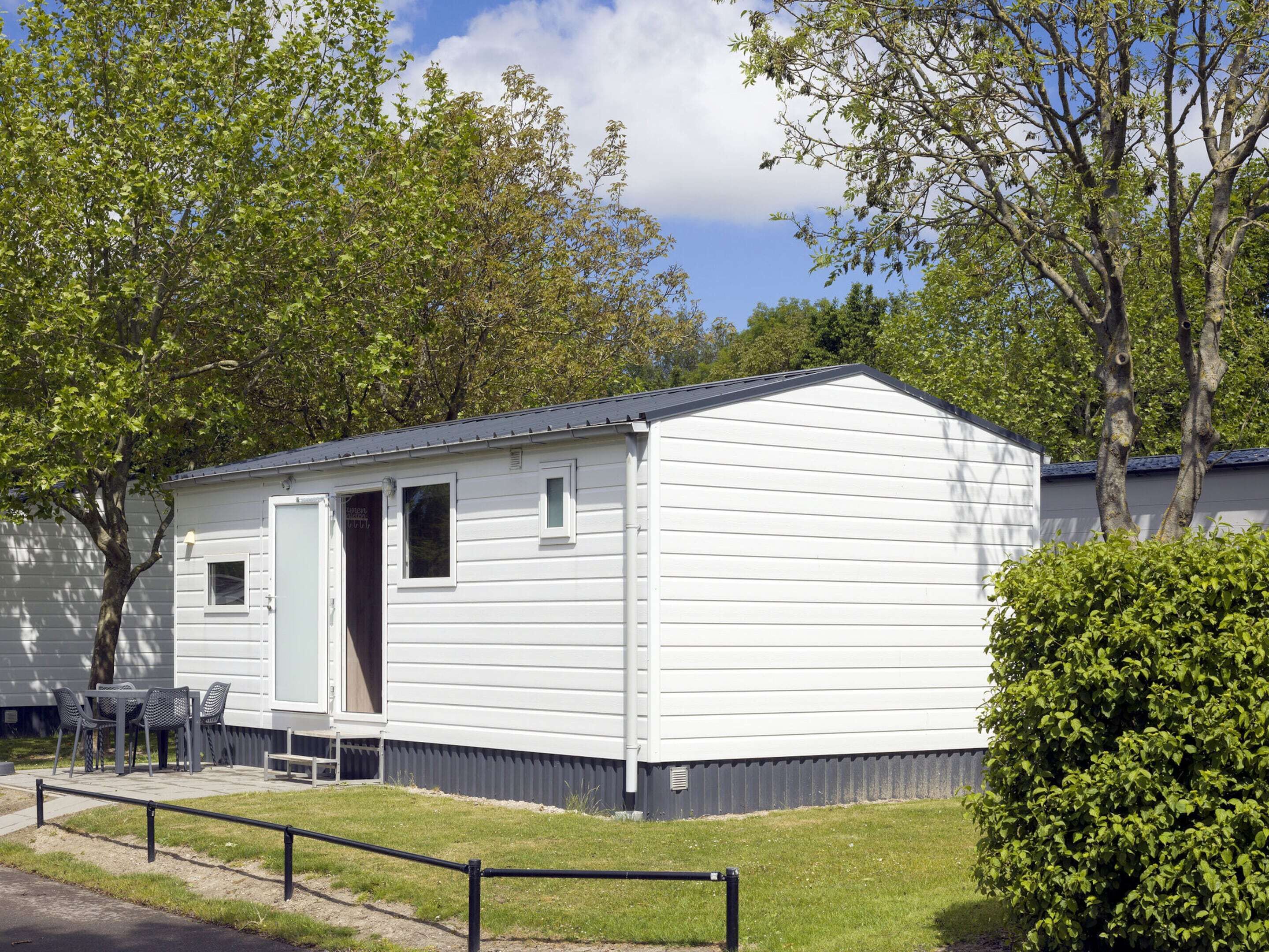 A single-story mobile home with a door open, a small porch with a table and chairs, and a grassy area in front.