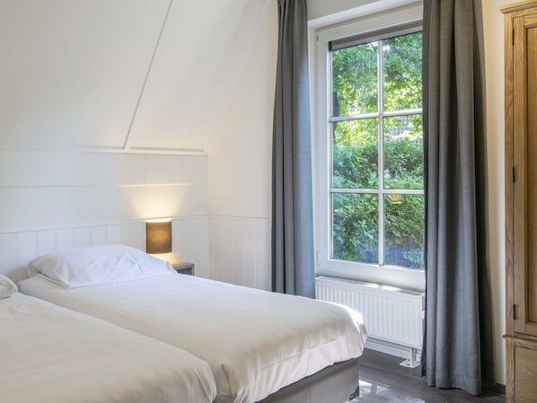 A bedroom with two beds, a nightstand with a lamp, a window with curtains, and a radiator.