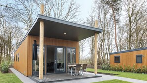 A modern wooden cabin with a covered patio, large sliding glass doors, and a small outdoor dining area. - Chic Chalet Near Almelo (Hoge Hexel)