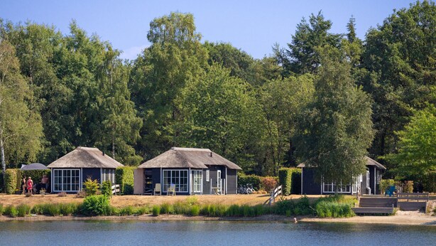 A lakeside property with thatched-roof cottages, a swimming area, and a forested backdrop. - Chic Chalet Near Almelo (Hoge Hexel)