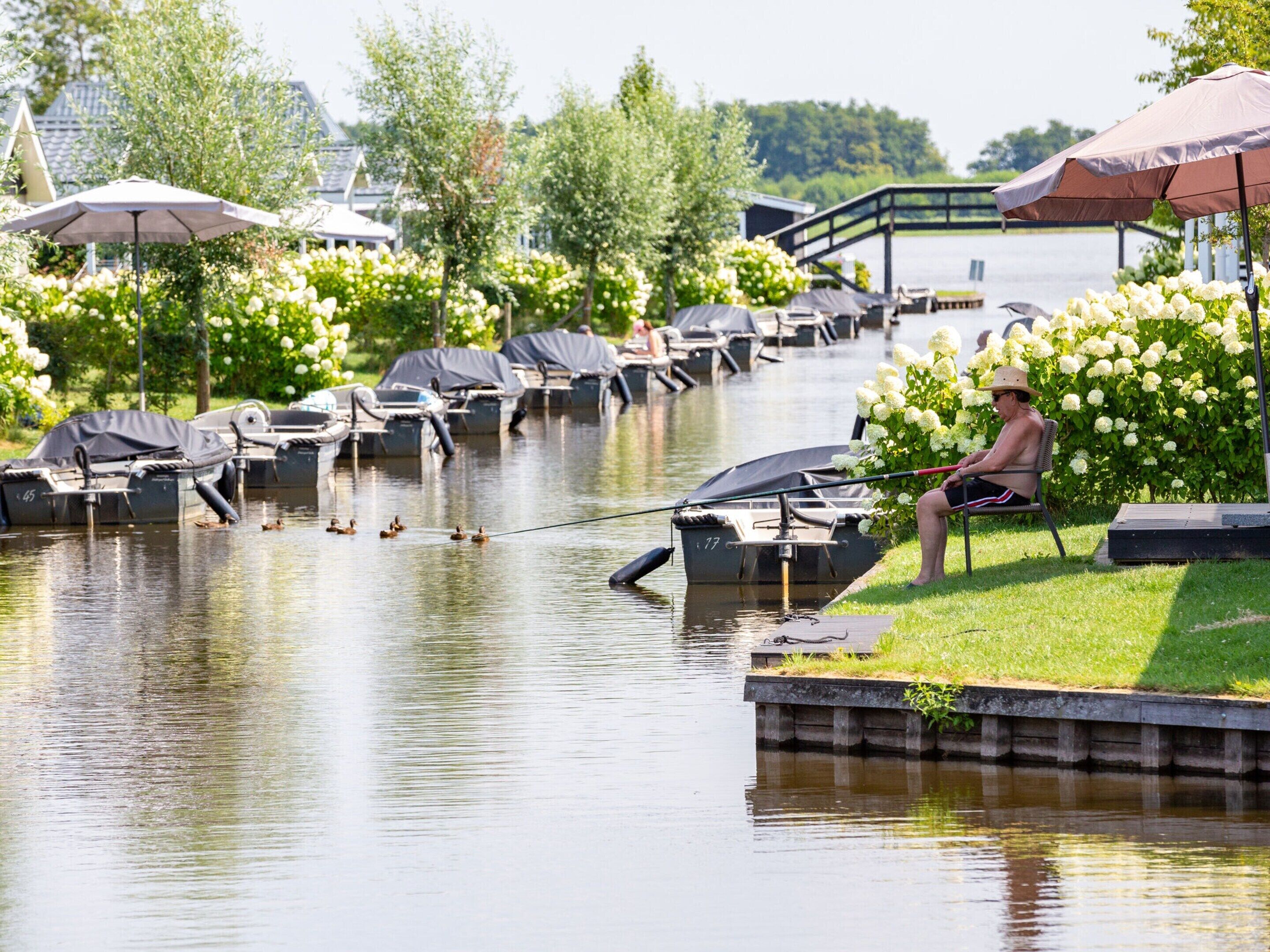 A riverside setting with boats docked, a person fishing, and a bridge in the background.