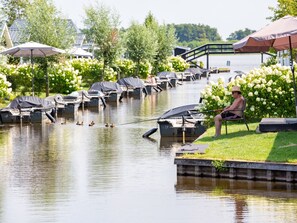 Un paysage au bord de la rivière, avec des bateaux amarrés, une personne qui pêche et un pont en arrière-plan.