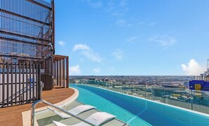 A rooftop infinity pool with a wooden deck and lounge chairs, overlooking a cityscape.