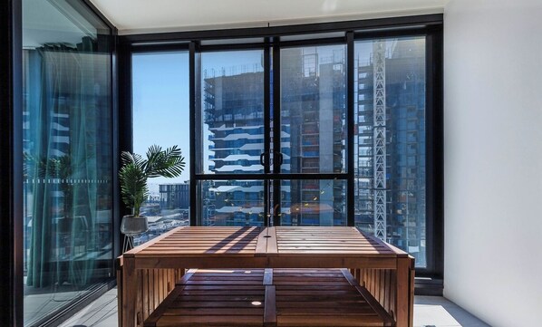 A modern balcony with a wooden table, large glass windows, and a potted plant.