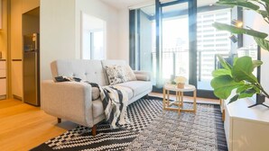 Un salon moderne avec un canapé, un tapis à motifs, une petite table d’appoint et une grande fenêtre donnant sur des immeubles.