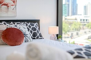 A bedroom with a bed, pillows, a nightstand with a lamp, and a window with a city view.